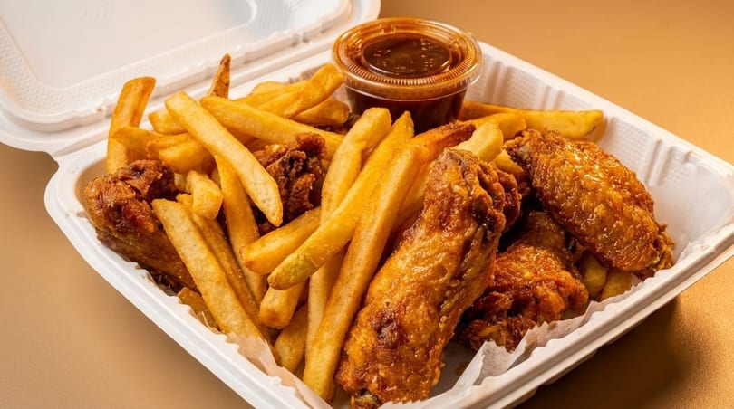Crispy fried chicken wings and golden french fries in a white takeout container with a side of sauce.