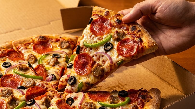 Hand lifting a slice of pepperoni, sausage, olive, onion, and bell pepper pizza from a box.